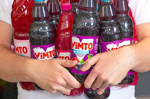 A man in a white T-shirt holding several different bottles of Vimto products
